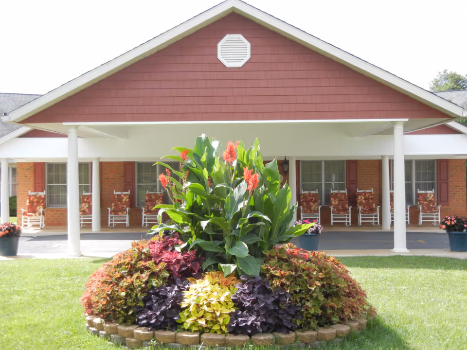 Front exterior view of a single-story assisted living facility with a covered porch featuring white columns and several rocking chairs with floral cushions. In front of the porch is a circular flower bed with various colorful plants and flowers, surrounded by a well-maintained lawn.