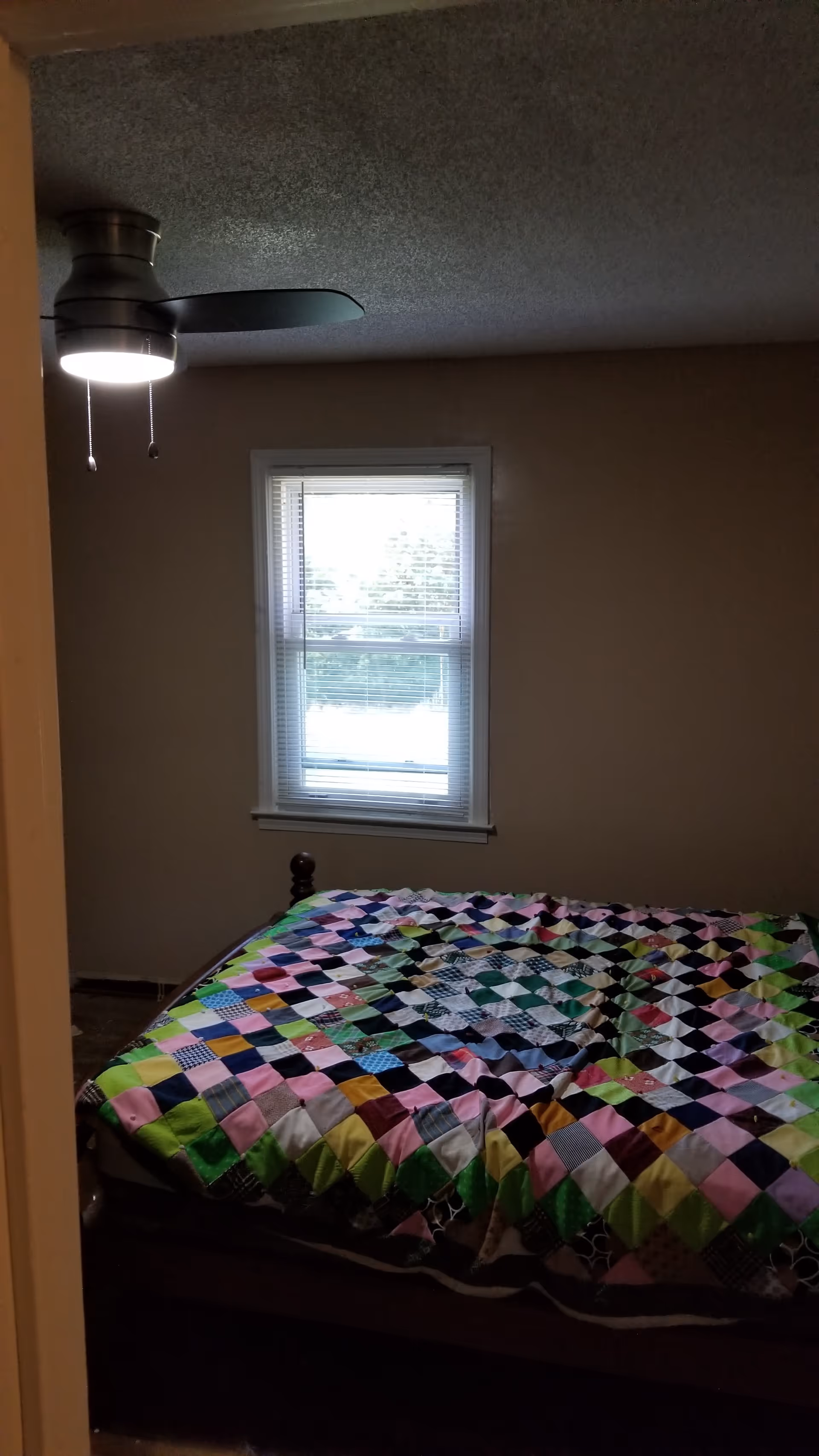 Bedroom with a colorful quilt on the bed, window with blinds, and a ceiling fan.