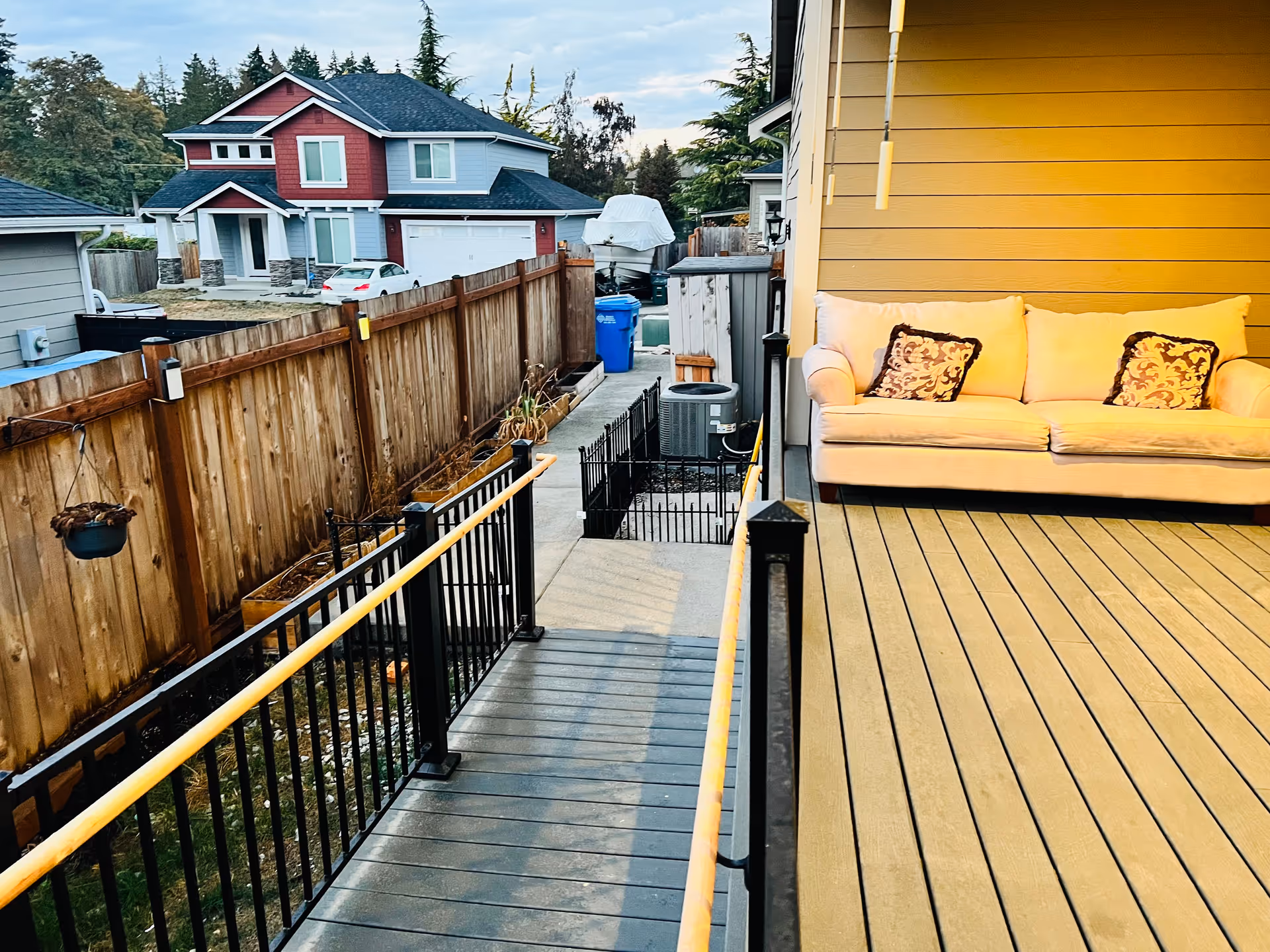 Outdoor wooden deck with sofa seating overlooks neighboring houses and a fenced yard.