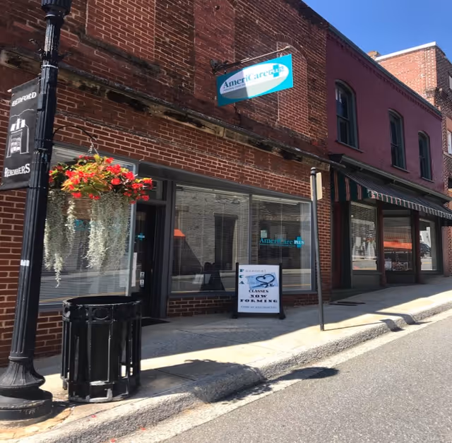 Brick storefront with AmeriCare Plus Home Care Bedford sign, hanging flower basket, and sidewalk.