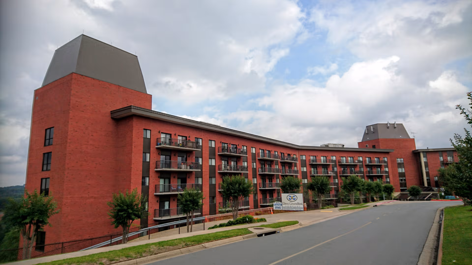 Red brick senior living building with balconies along a curved facade and a driveway in front.