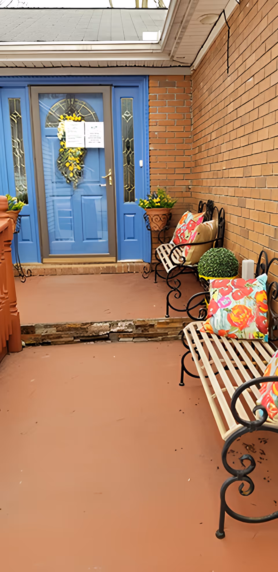 Covered brick entryway with a bright blue front door, a wreath on the door, benches with colorful floral cushions, and potted plants.