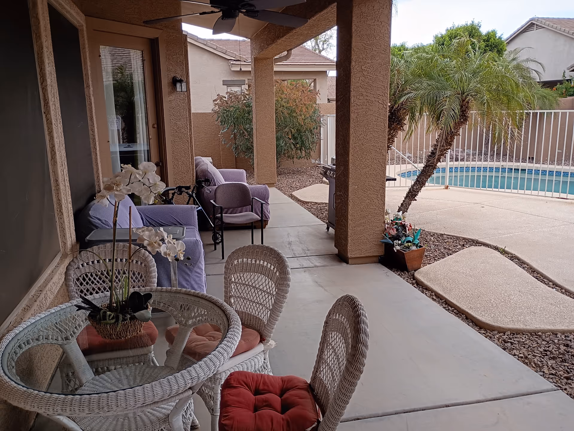 Covered patio with wicker seating, cushions, potted plants, and a pool behind a white fence.