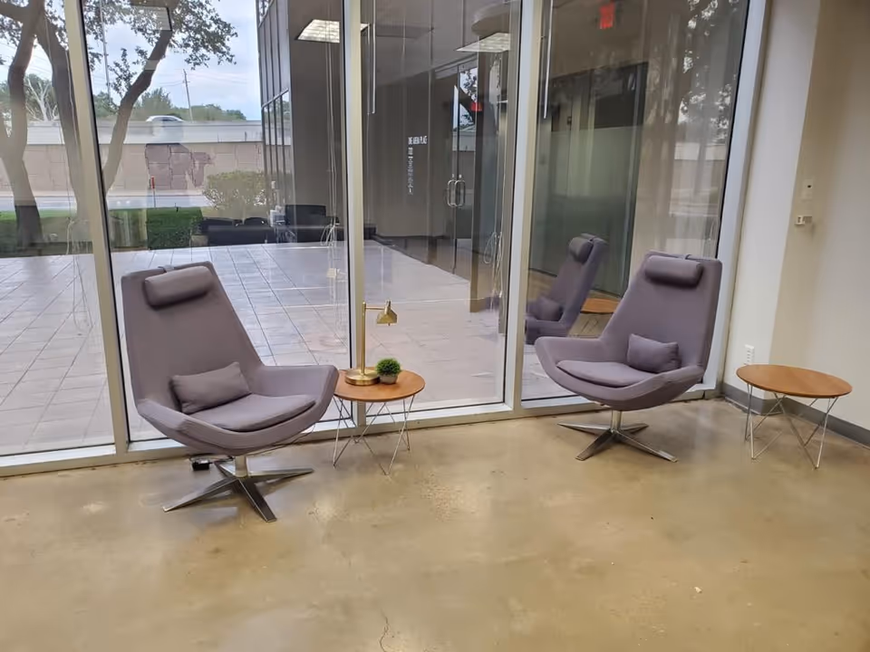 Two lavender lounge chairs with small tables sit in a modern glass-walled lounge area.
