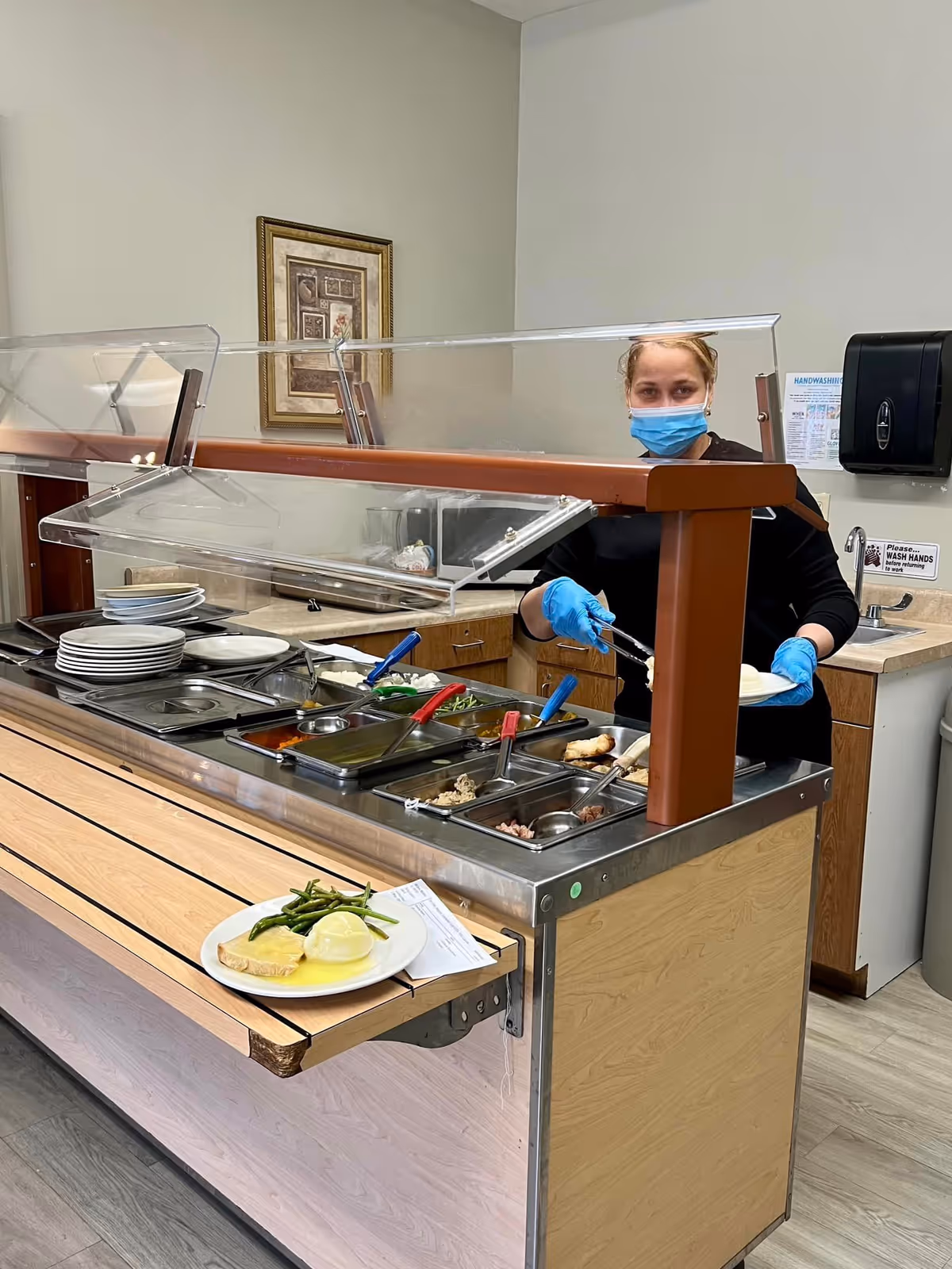 Staff member in mask serves food at a cafeteria dining line in a senior living facility.
