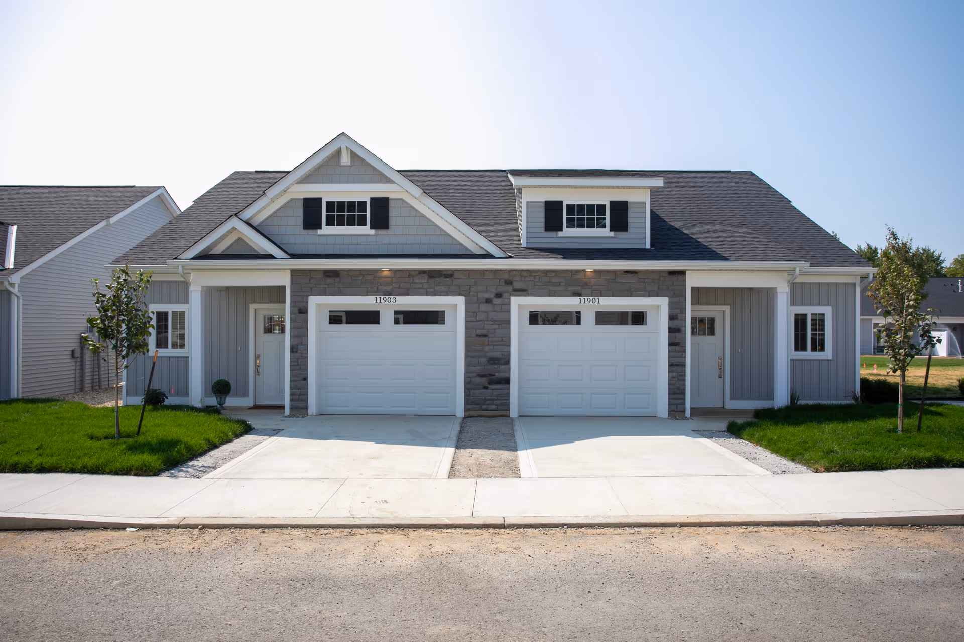 Front exterior view of a modern duplex building with two white garage doors, two front doors, small windows, and a well-maintained lawn with young trees on either side.