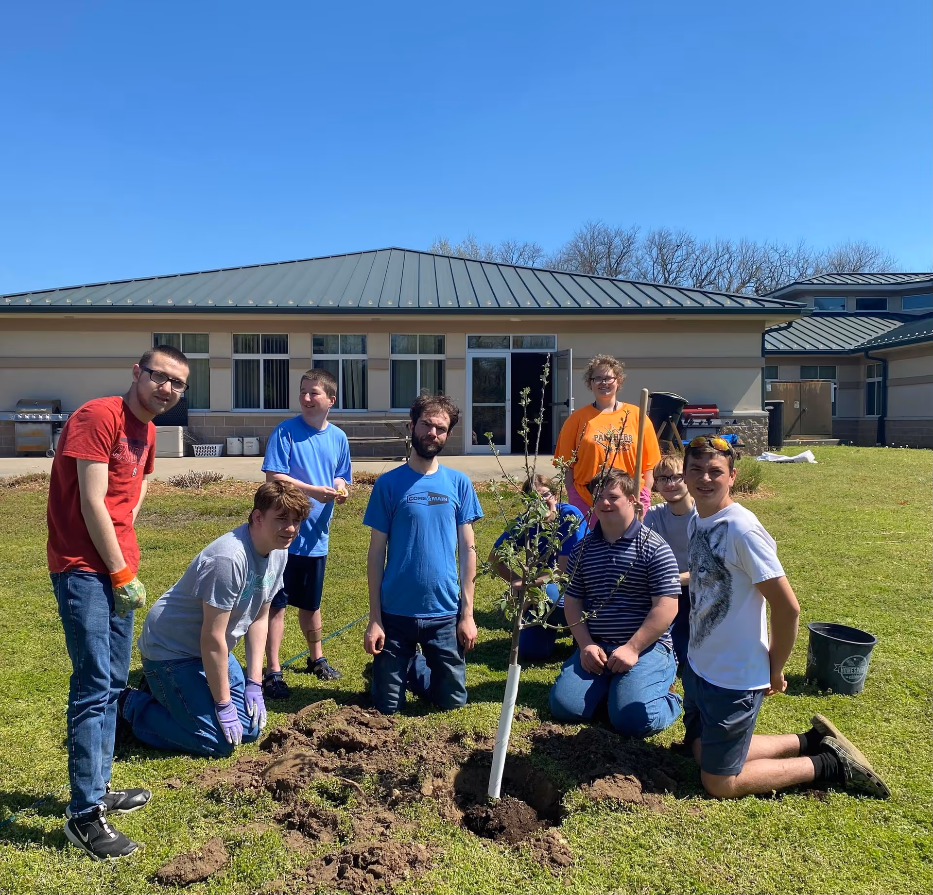 Group of volunteers planting a tree on a sunny lawn in front of a facility.