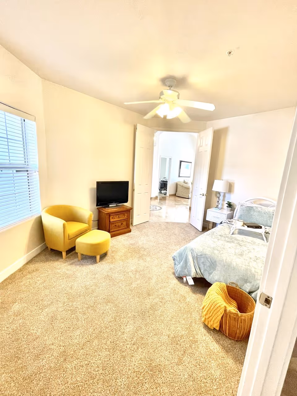 Bright furnished bedroom with a bed and nightstand, TV on a wooden dresser, a yellow armchair with ottoman, and a woven basket on carpeted floor.