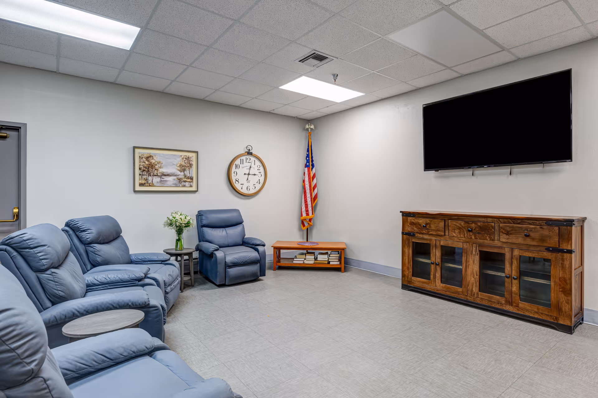 Bright senior living lounge with blue recliners, wall clock, American flag, TV, and cabinet.