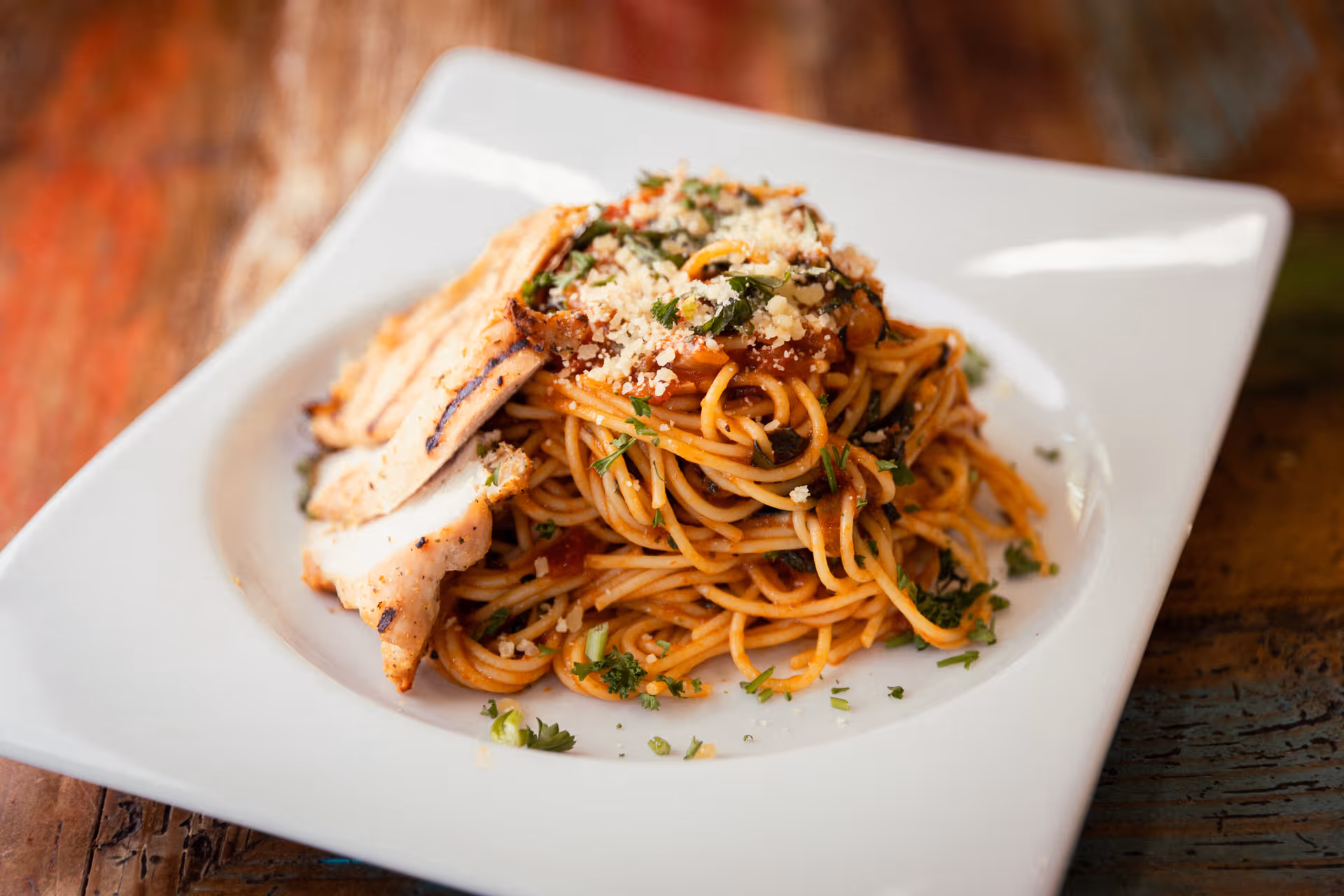 A white square plate with a serving of spaghetti topped with tomato sauce, grated cheese, chopped herbs, and slices of grilled chicken breast on a wooden table.