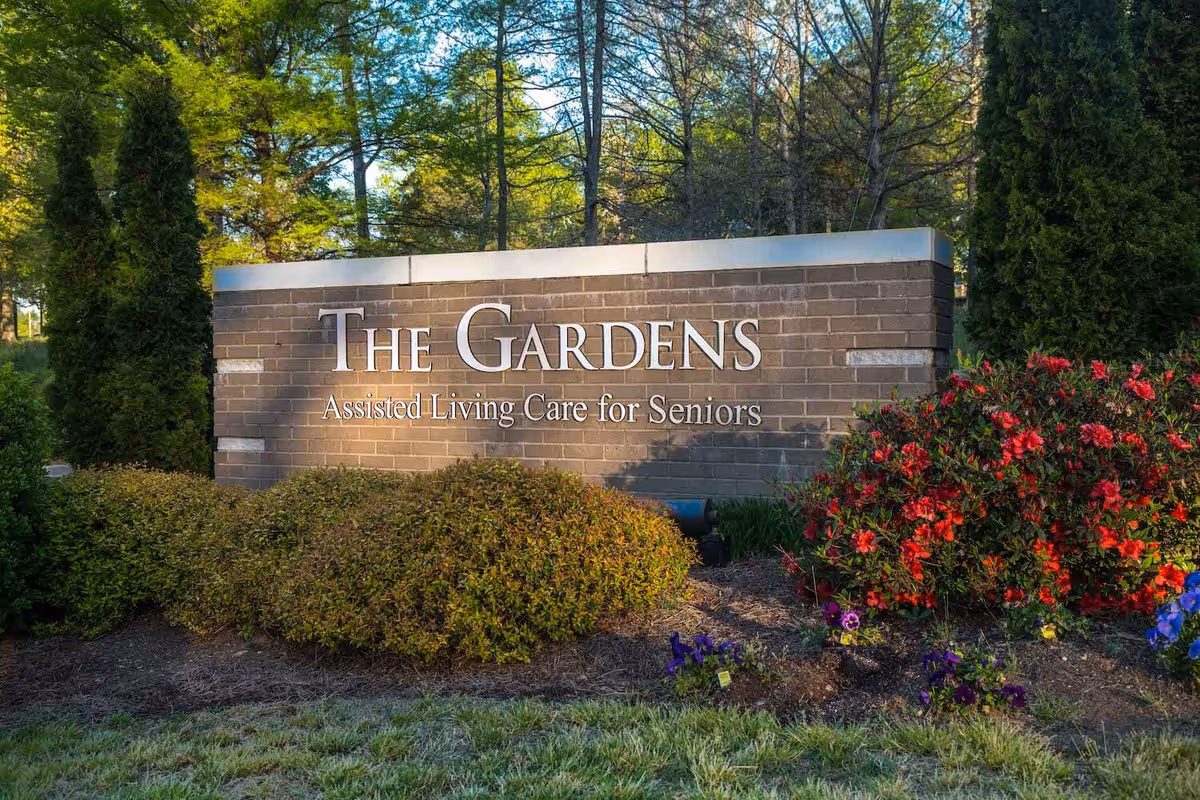 Brick entrance sign reading "The Gardens Assisted Living Care for Seniors" surrounded by shrubs, flowers, and trees.