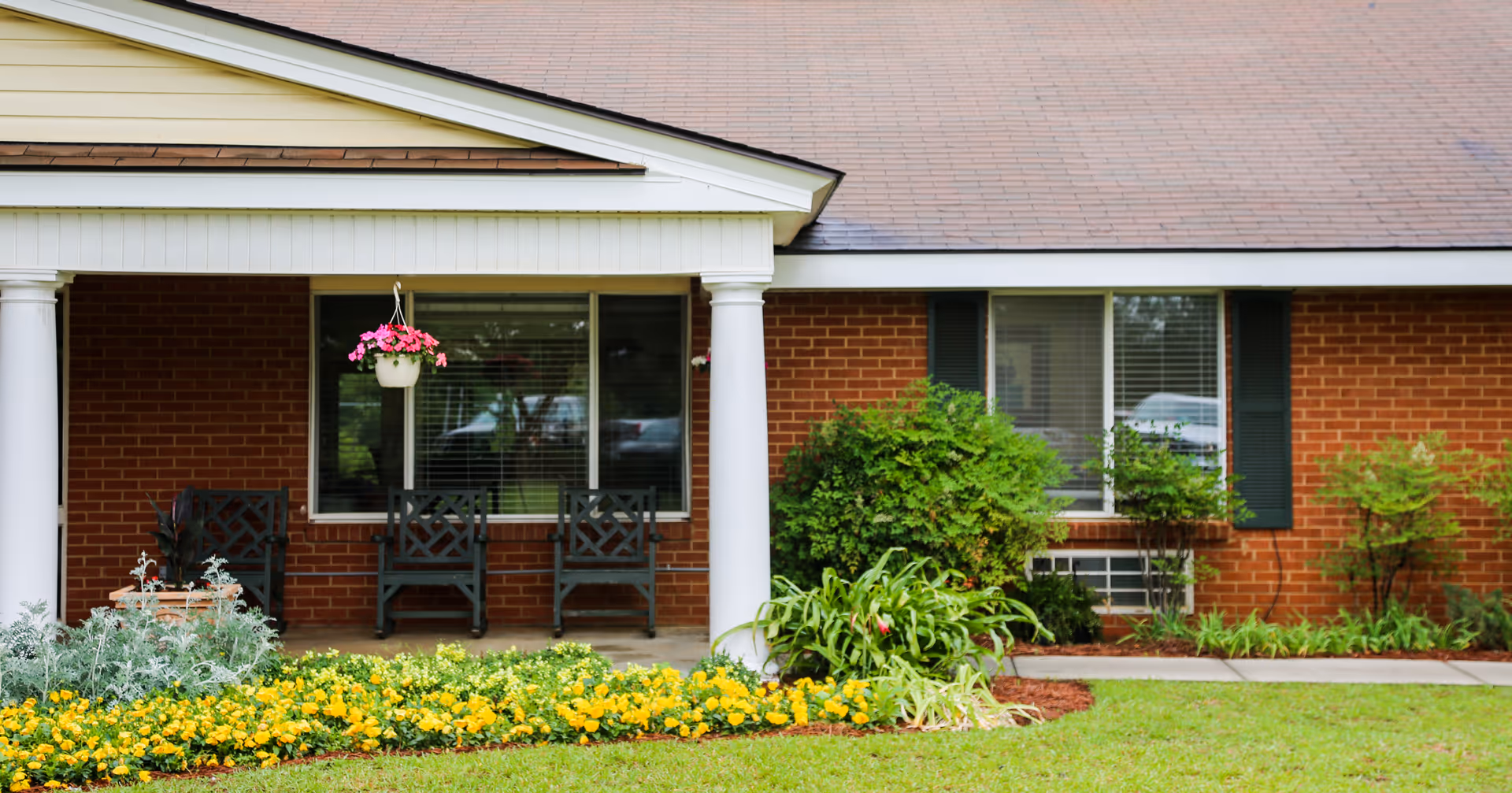 Front porch of a brick building with white columns, three black chairs, a hanging pot with pink flowers, and a garden bed with yellow flowers and green shrubs in front.