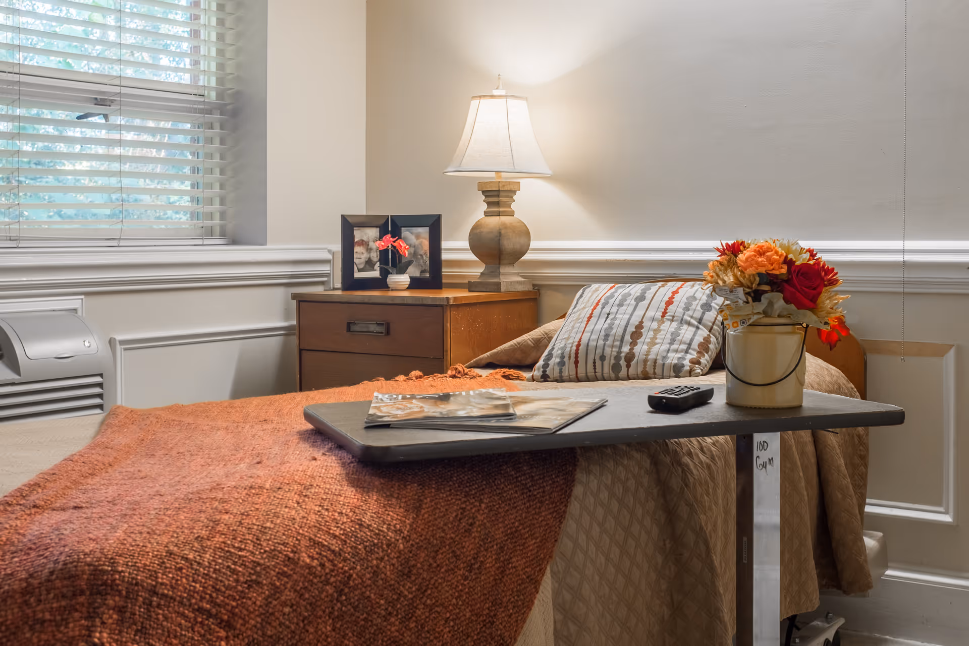 A neatly made bedroom in a senior living facility with a bed, lamp, tray, and flowers.