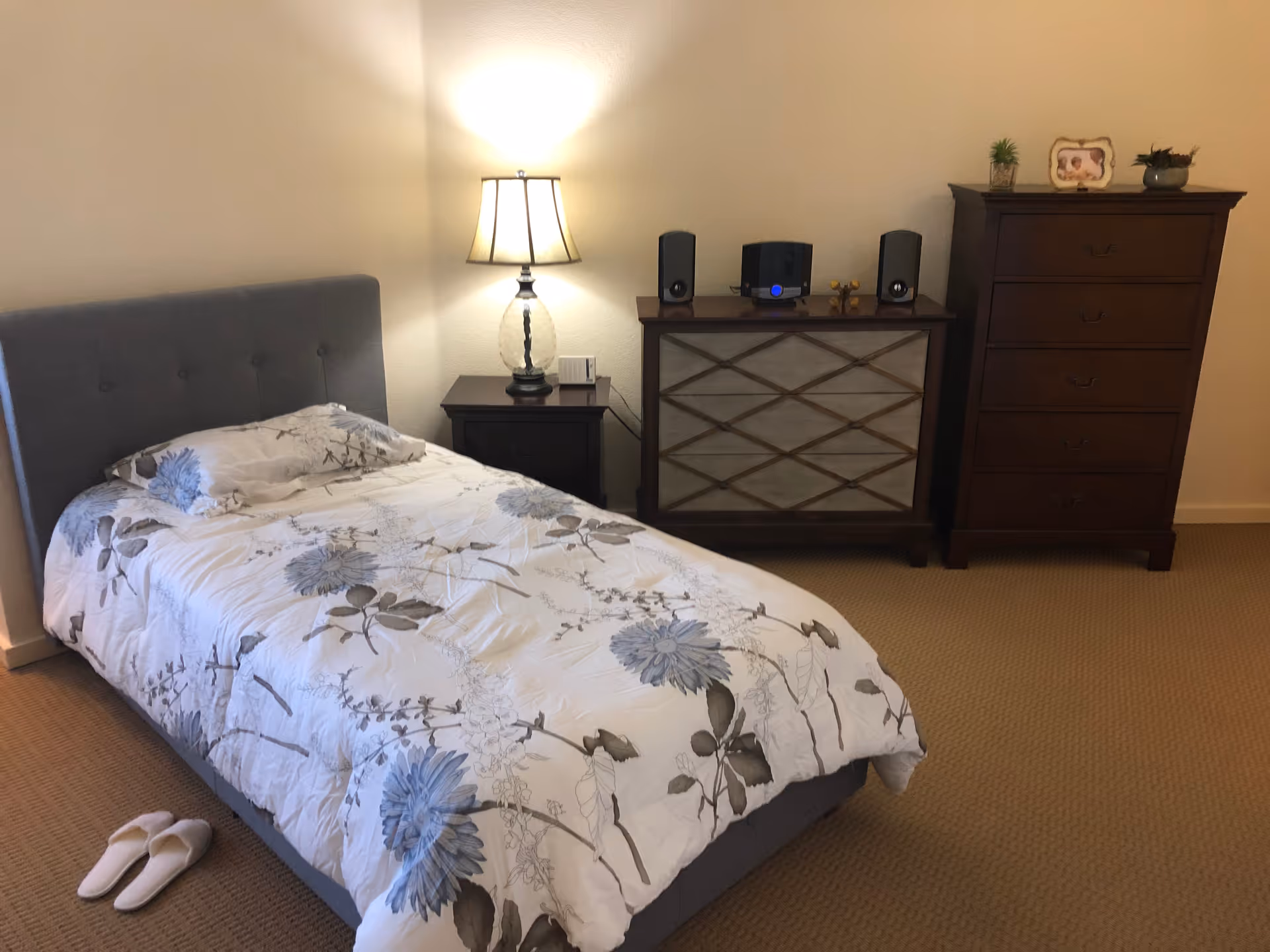 Cozy bedroom with a patterned bed, nightstand lamp, dresser, and slippers on carpet.