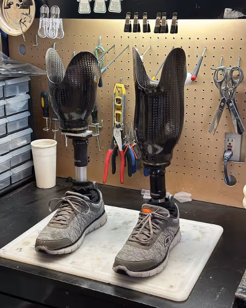 Two prosthetic legs wearing athletic shoes stand on a workbench in a tool-filled workshop.