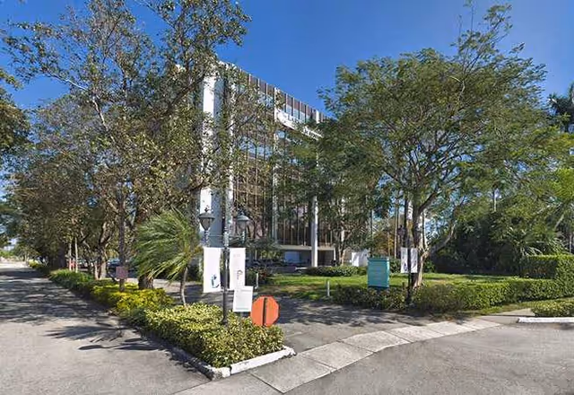 Modern glass-front senior living building surrounded by trees and manicured landscaping.