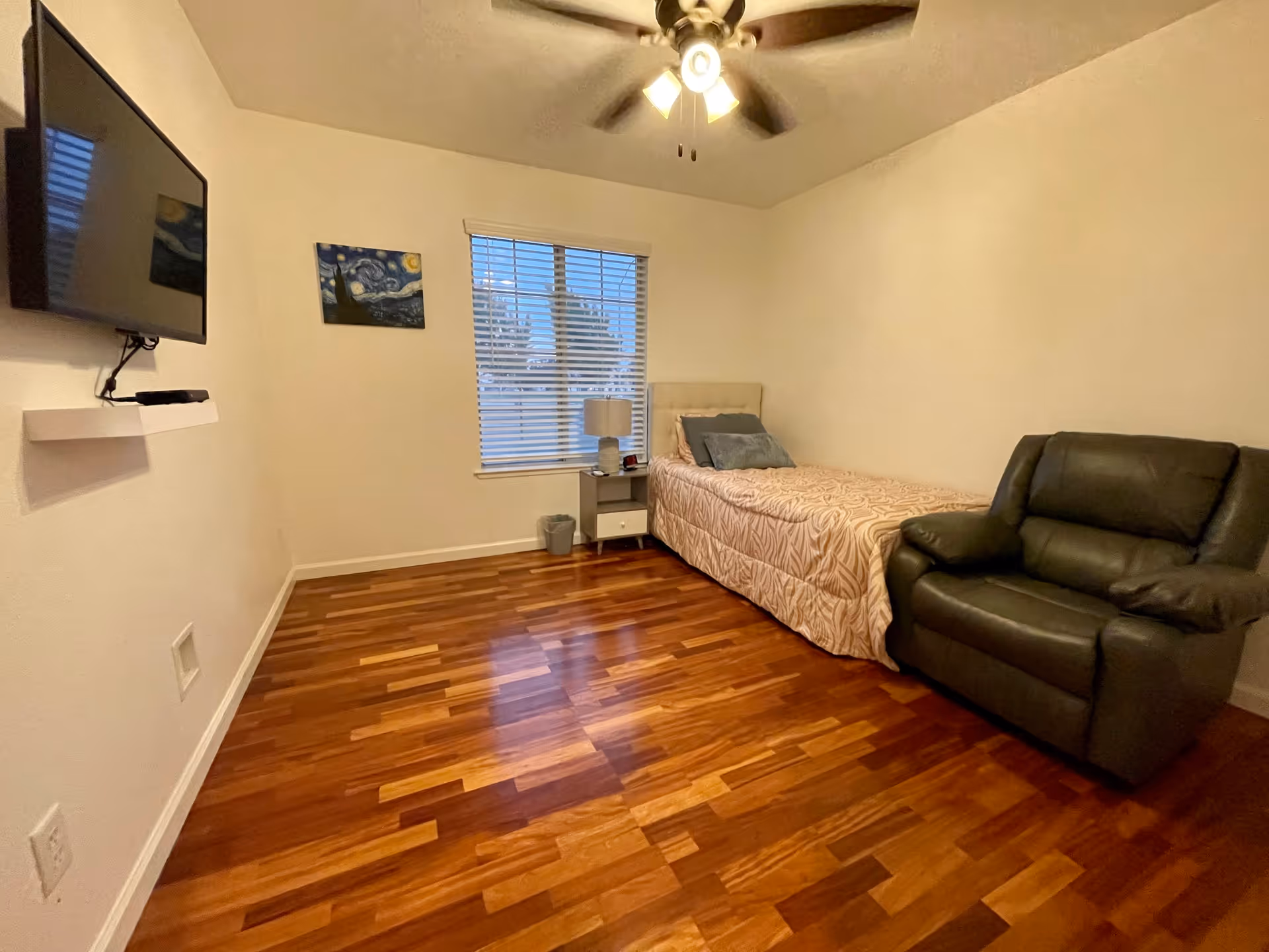 A small bedroom with wooden flooring, a single bed with a beige patterned bedspread, a gray armchair, a nightstand with a lamp and an alarm clock, a window with blinds, a ceiling fan with lights, a wall-mounted TV, and a small painting on the wall.