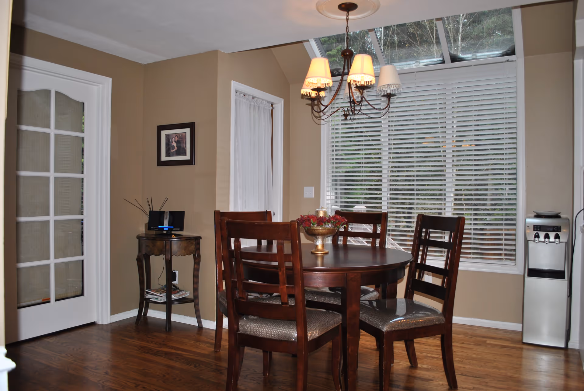 Dining area with a round wooden table and four chairs under a chandelier next to a large window with blinds.