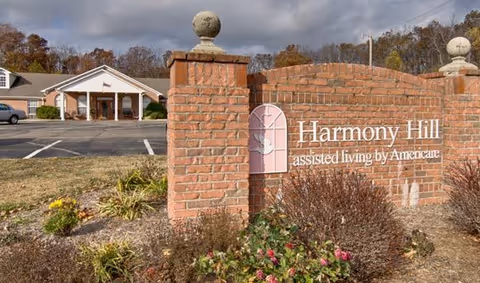 Brick entrance sign for 'Harmony Hill assisted living by Americare' with the facility building and parking lot in the background.