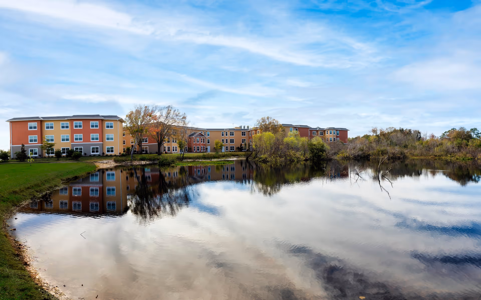 A large senior living facility building with multiple floors and colorful exterior walls in shades of orange, yellow, and red, reflected in a calm pond in the foreground. Trees and greenery surround the building and pond under a partly cloudy blue sky.