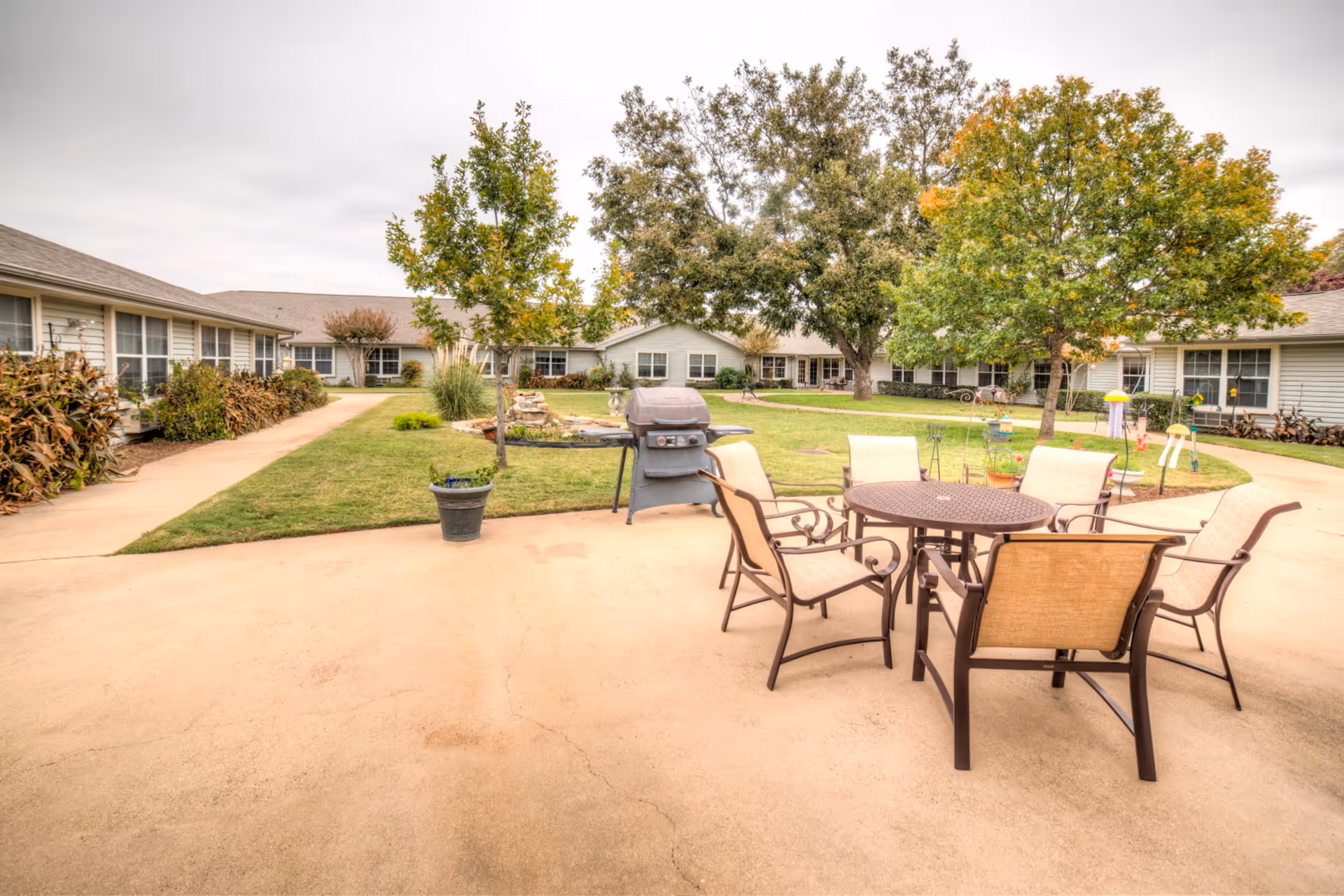 A central courtyard at an assisted living facility with a patio table and chairs, a grill, lawn, trees, and surrounding single-story buildings.