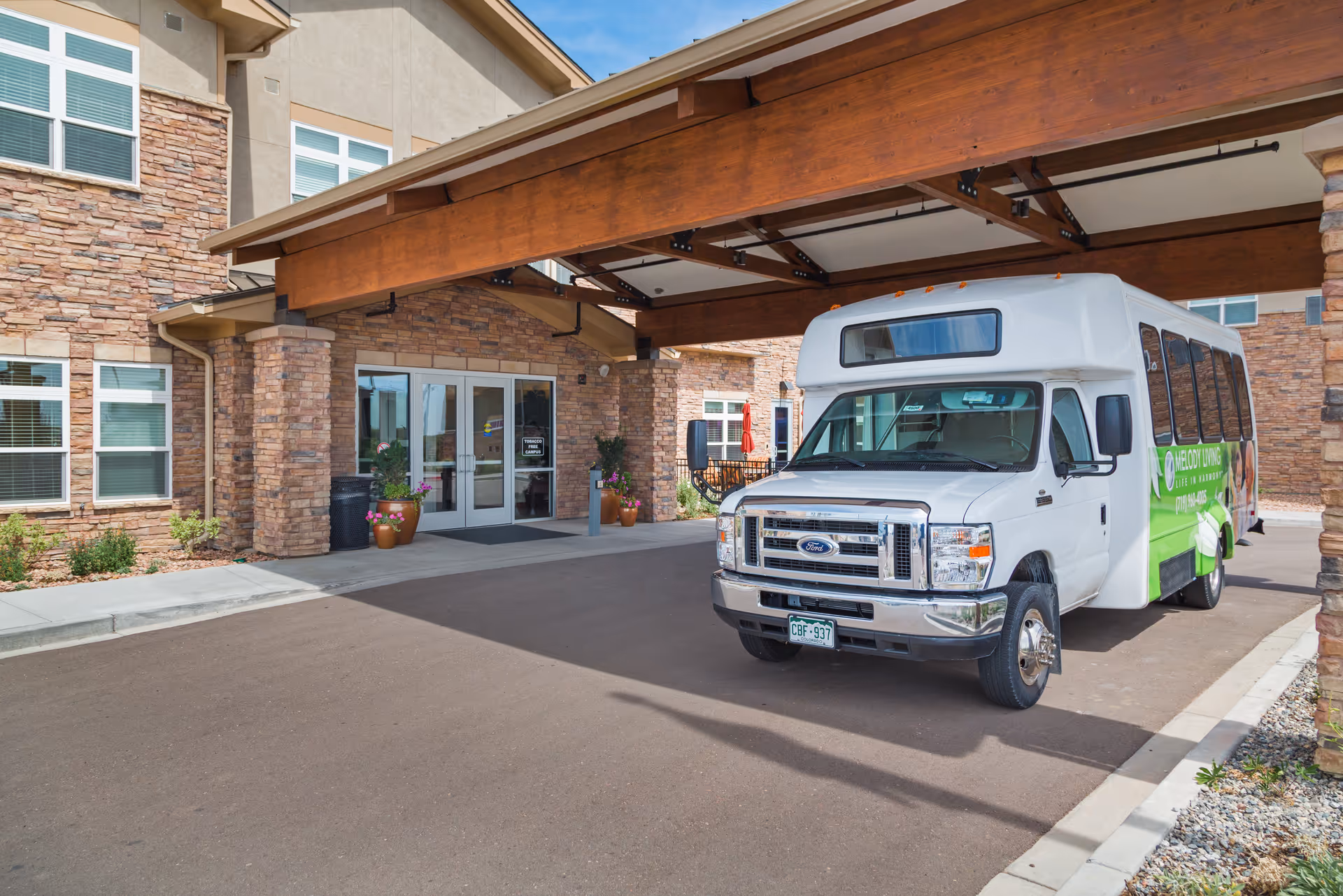 Exterior view of Melody Living facility entrance with a covered drop-off area and a white shuttle bus parked in front. The building has stone and beige siding with multiple windows and potted plants near the entrance doors.