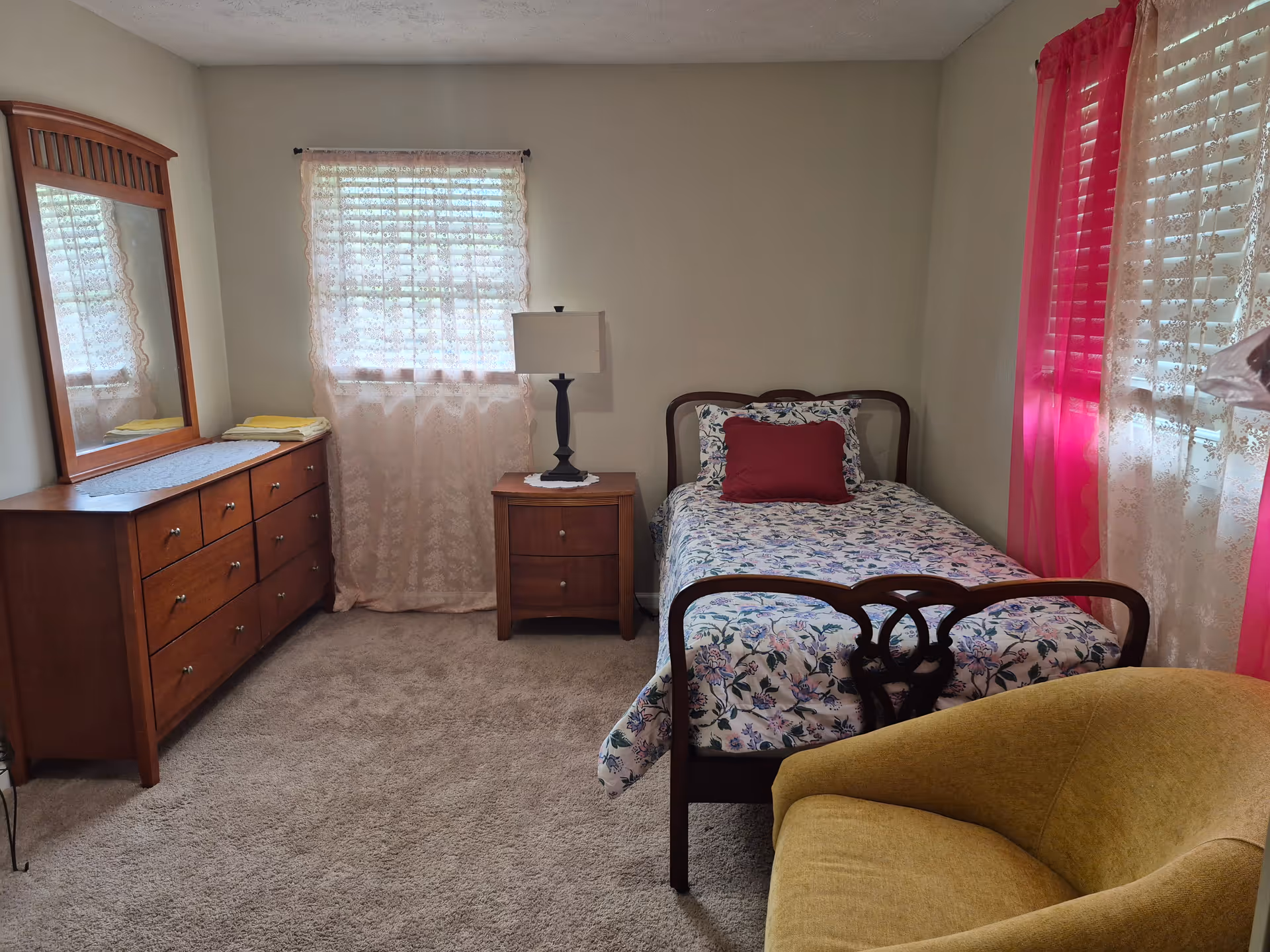 Cozy single bedroom with floral bedspread, wooden dresser, nightstand, lamp, and pink sheer curtains.