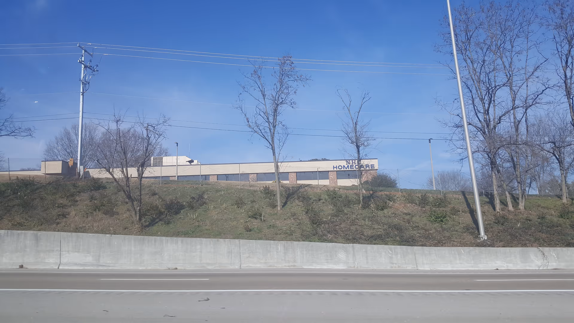 Low, single-story building labeled NHC HomeCare on a hillside beside a highway.