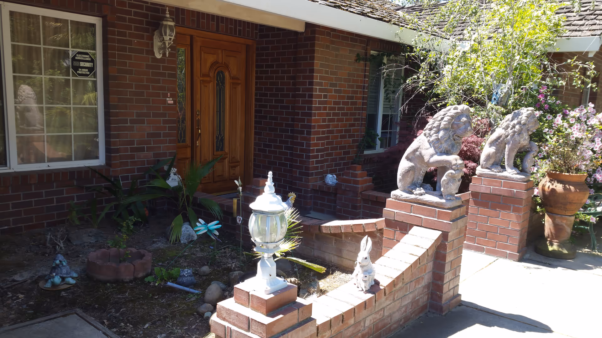 Front entrance of a brick building with a wooden double door, two stone lion statues on brick pillars, a small garden area with plants and decorative items including a white lamp and a rabbit statue, and a window with a security sign.