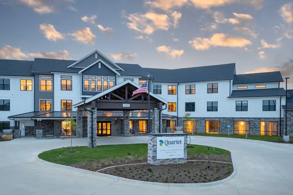 Exterior front entrance of Quartet senior living building with a covered porte-cochere, American flag, illuminated windows, and a landscaped sign in the foreground.