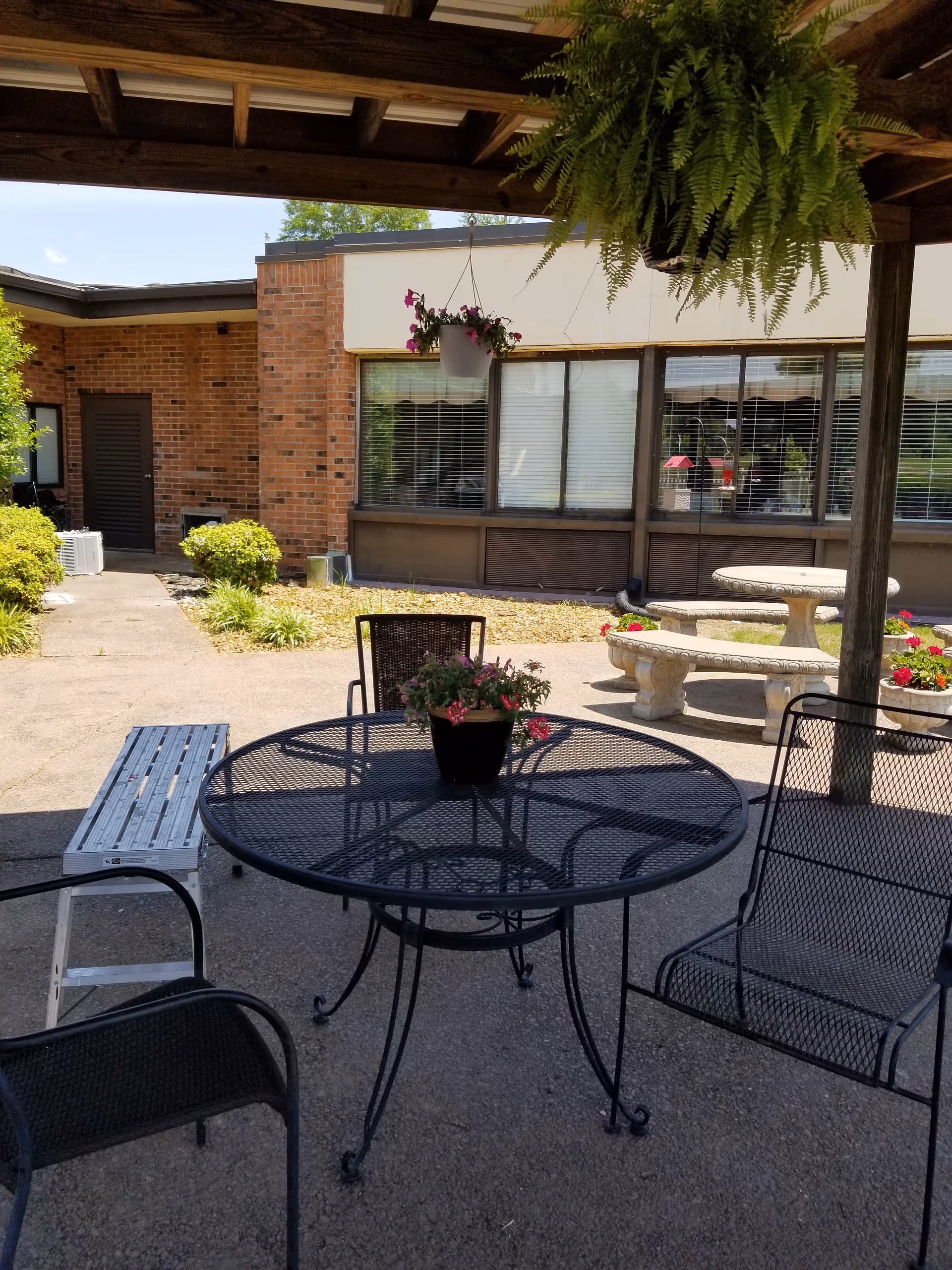 Outdoor patio with metal table, chairs, hanging fern, and brick building facade.