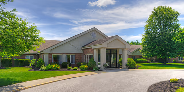 Front exterior of a single-story nursing home building with a covered entrance, landscaped lawn, and trees under a blue sky.