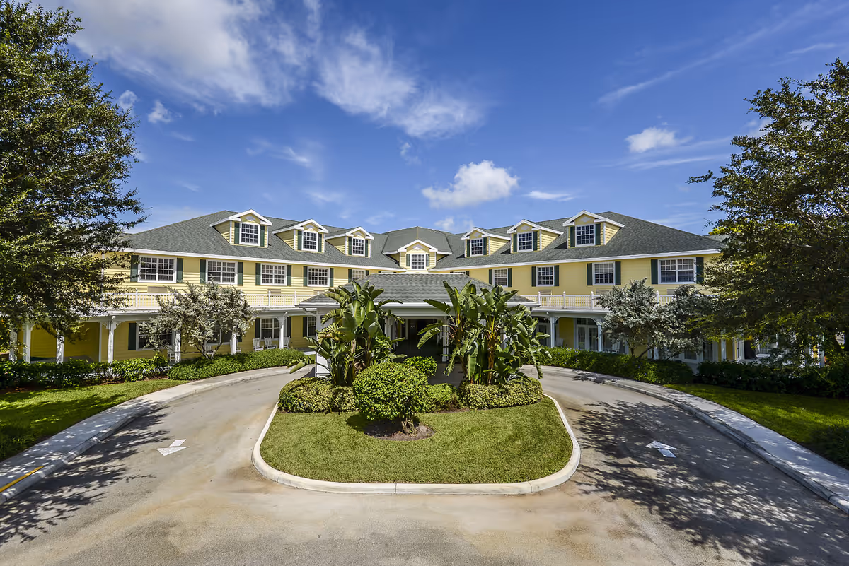 Yellow three-story senior living building with a covered central entrance, circular driveway and landscaped greenery under a blue sky.
