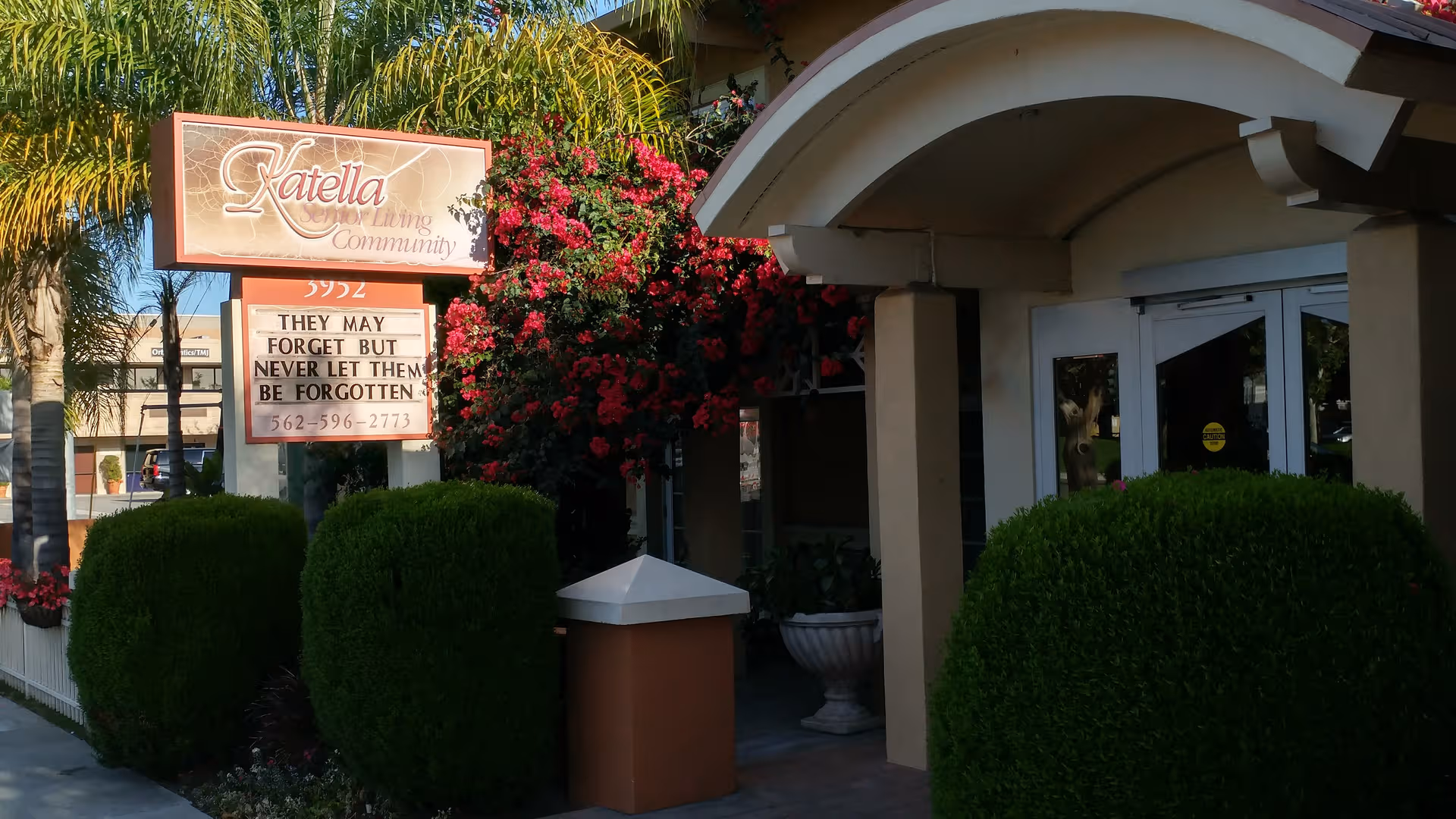 Exterior view of Katella Senior Living Community entrance with manicured hedges, palm trees, and bougainvillea
