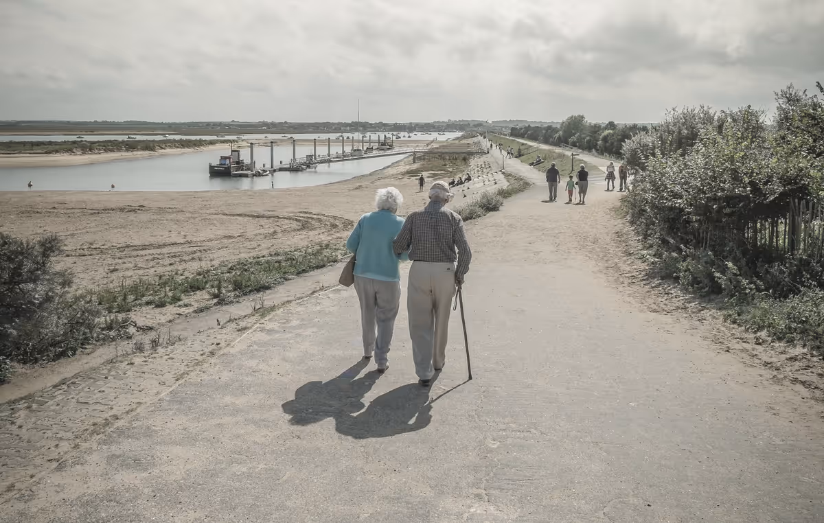 Elderly couple walks along a riverside path with other pedestrians.