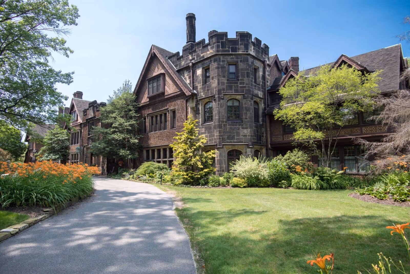 Exterior view of a large, historic-style brick building with a stone turret, surrounded by well-maintained green lawns, trees, and orange flowers under a clear blue sky.