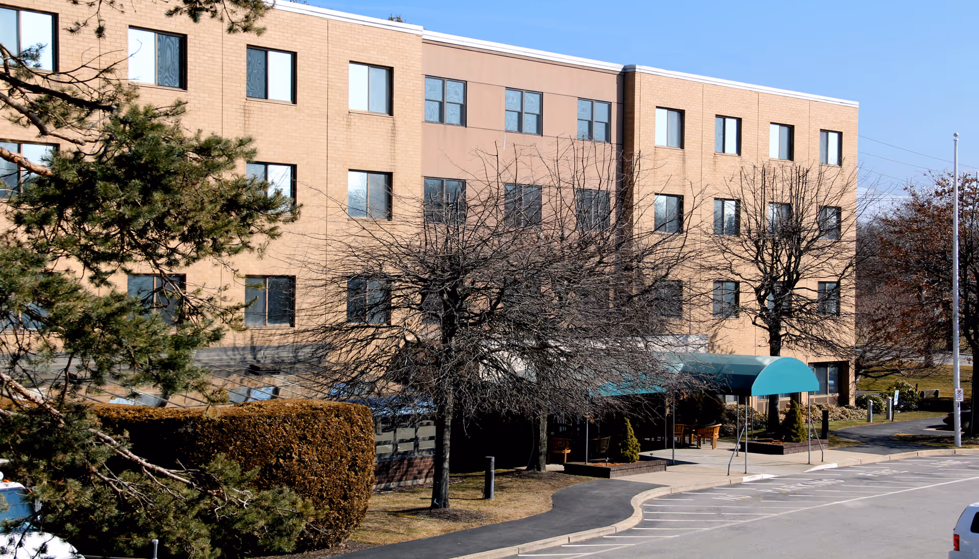 Exterior view of a three-story brick building with multiple windows, a green awning over the entrance, leafless trees, and a parking area in front under a clear blue sky.