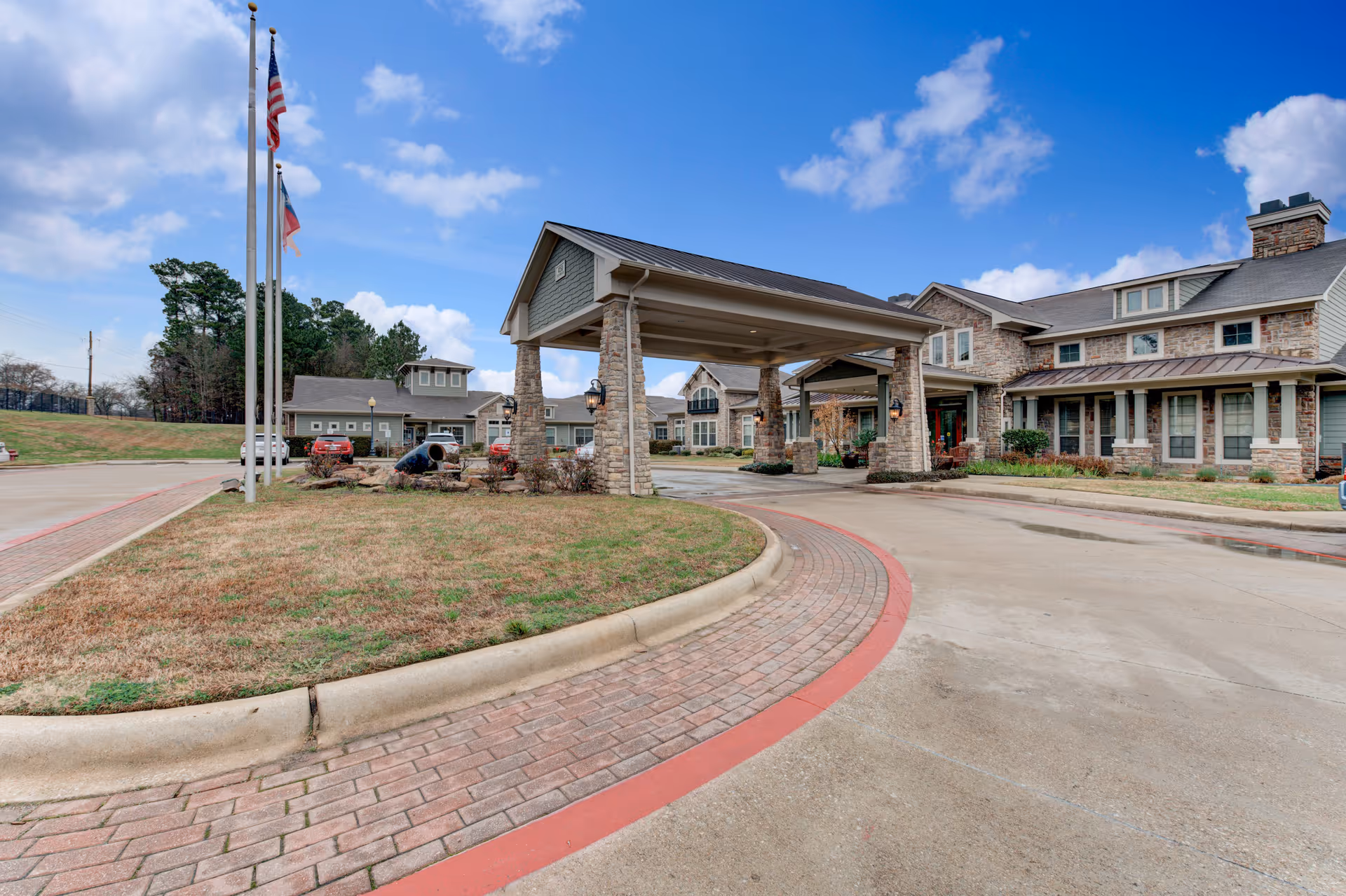 Stone-front entrance with porte-cochère, flags, and curved brick driveway at a senior care facility.