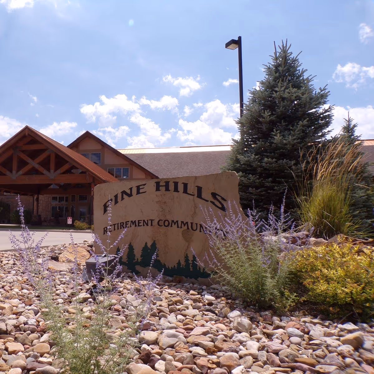 Stone sign reading 'Pine Hills Retirement Community' sits among rocks and plants with the facility entrance and evergreen trees behind it.