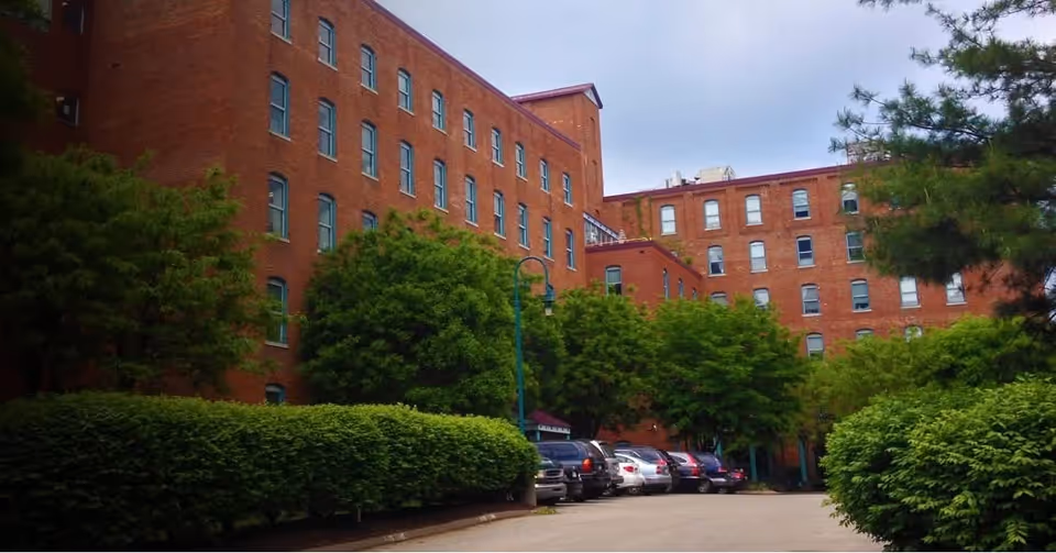 Brick senior-living building exterior with parked cars and lush green trees.