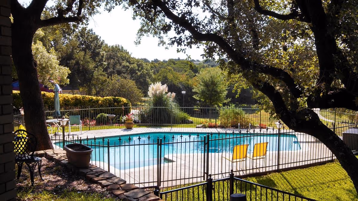 Fenced outdoor swimming pool with two yellow lounge chairs, patio furniture, potted plants, and large trees overlooking a landscaped yard.