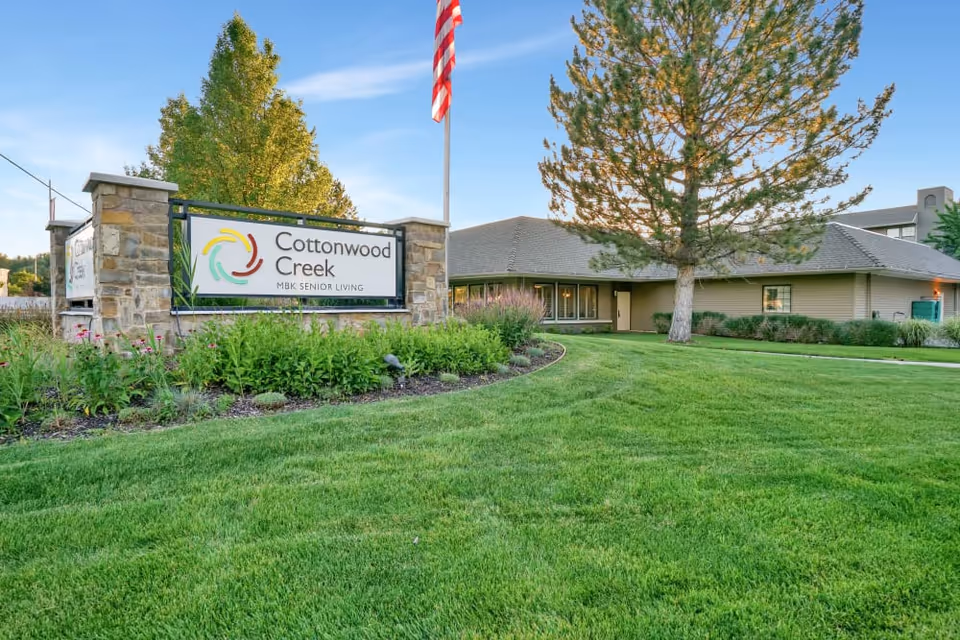 Sign for Cottonwood Creek MBK Senior Living on a stone base with a manicured lawn, the facility building, and an American flag in the background.