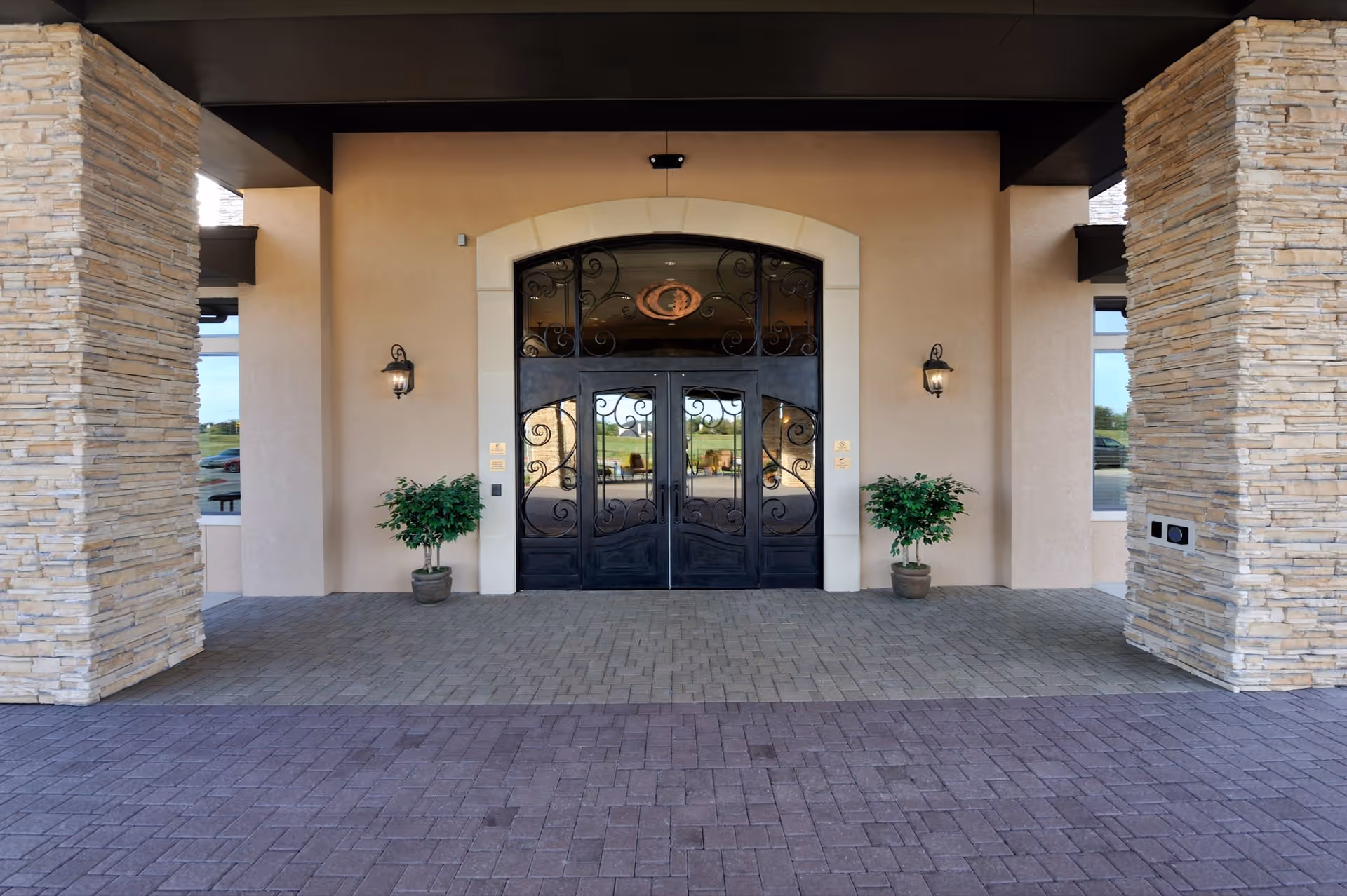 Entrance to a building with double black wrought iron doors framed by a light stone arch. Two potted plants flank the doors, and two wall-mounted lantern-style lights are on either side. The entrance is covered by a roof supported by stone pillars, and the ground is paved with bricks.