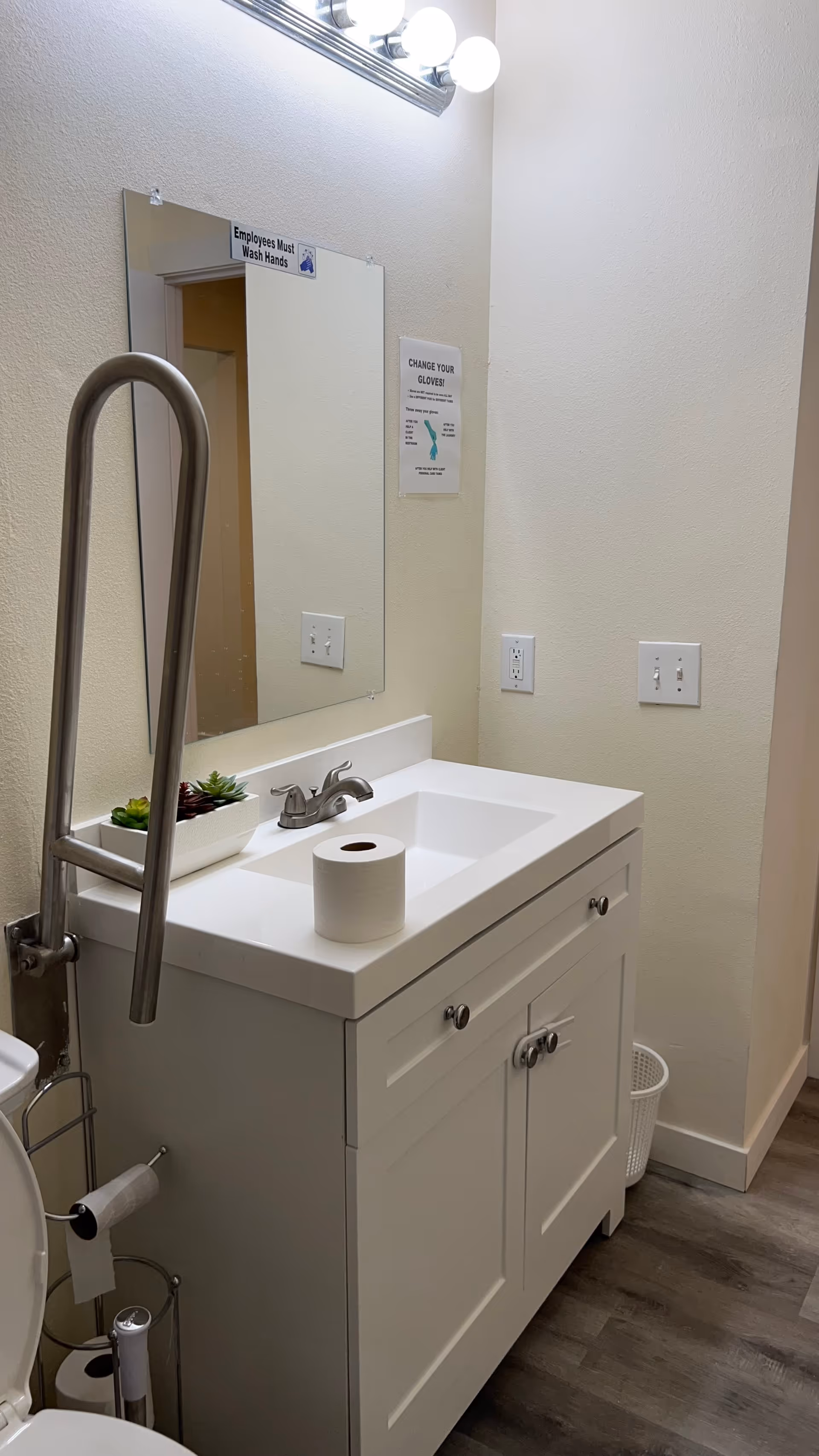 Accessible bathroom with sink vanity, mirror, handrail, and hand hygiene posters.