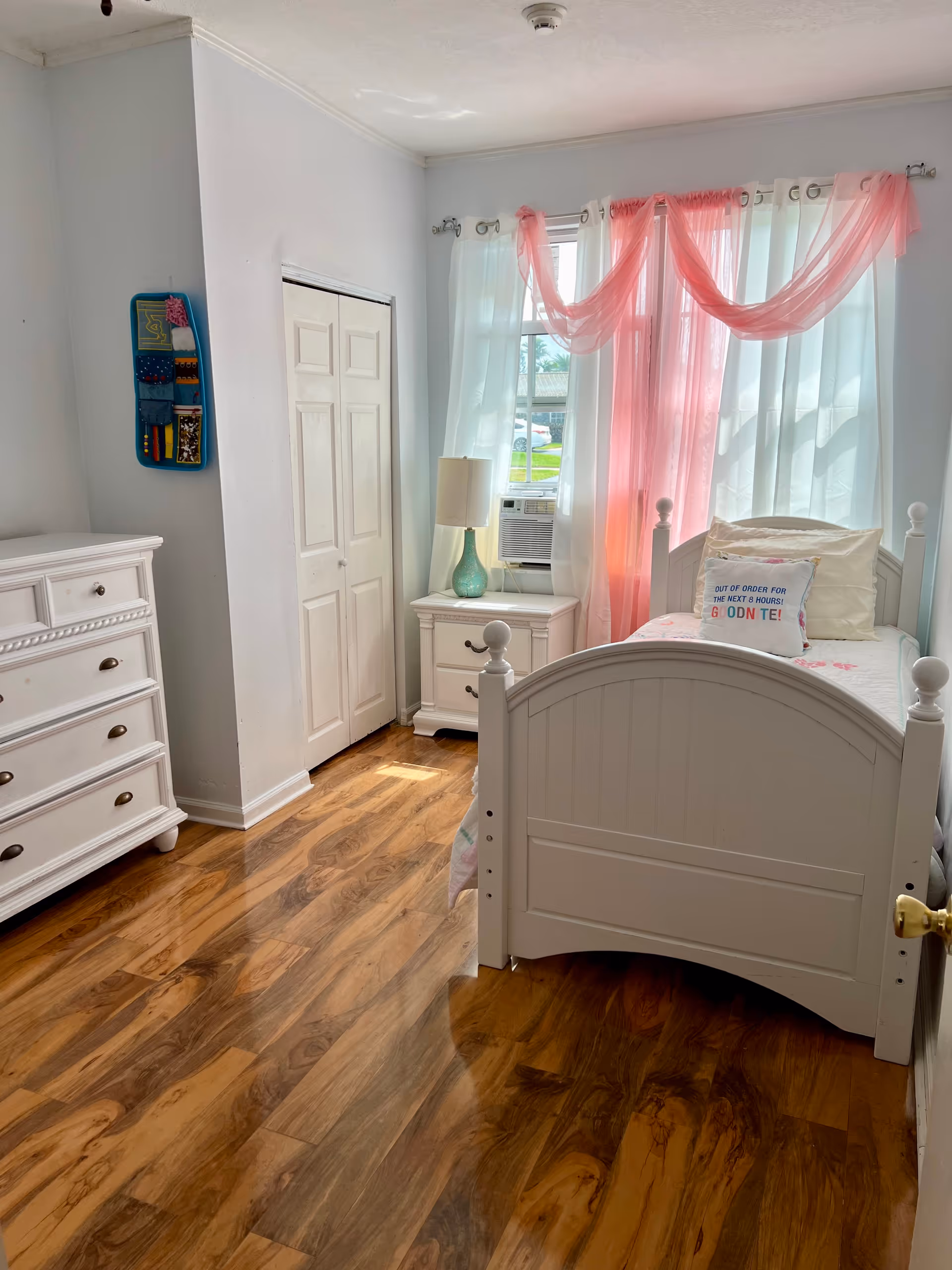 Bright bedroom with white furniture, pink curtains, and wood flooring.