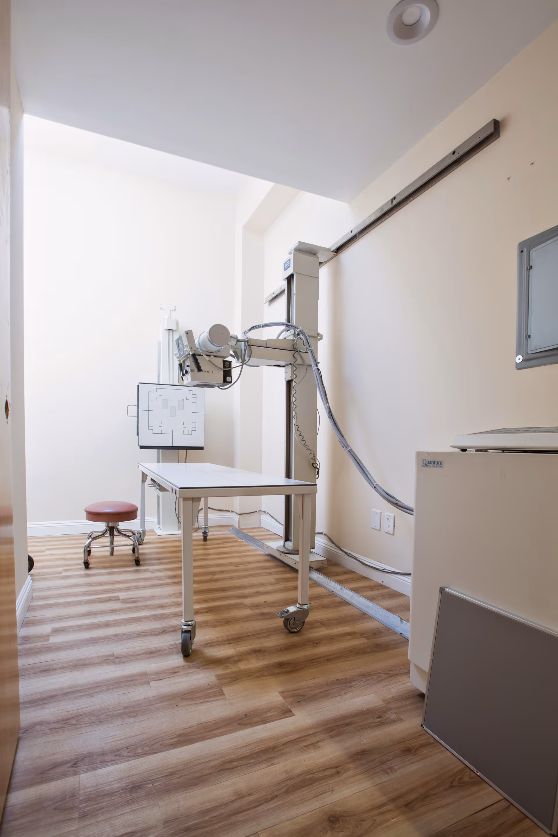 An X-ray imaging room with a vertical radiography machine, a wheeled table, and a red stool.