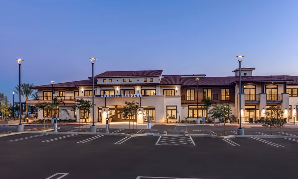 Front exterior of a two-story Merrill Gardens senior living building at dusk with illuminated entrance and an empty parking lot.