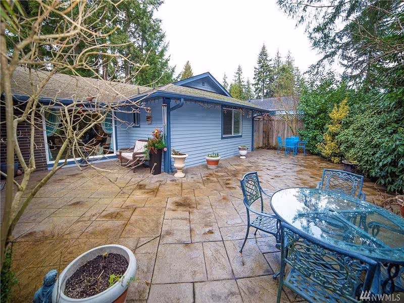 Outdoor patio area with a glass-top metal table and chairs, potted plants, and a blue building with windows surrounded by trees and greenery.