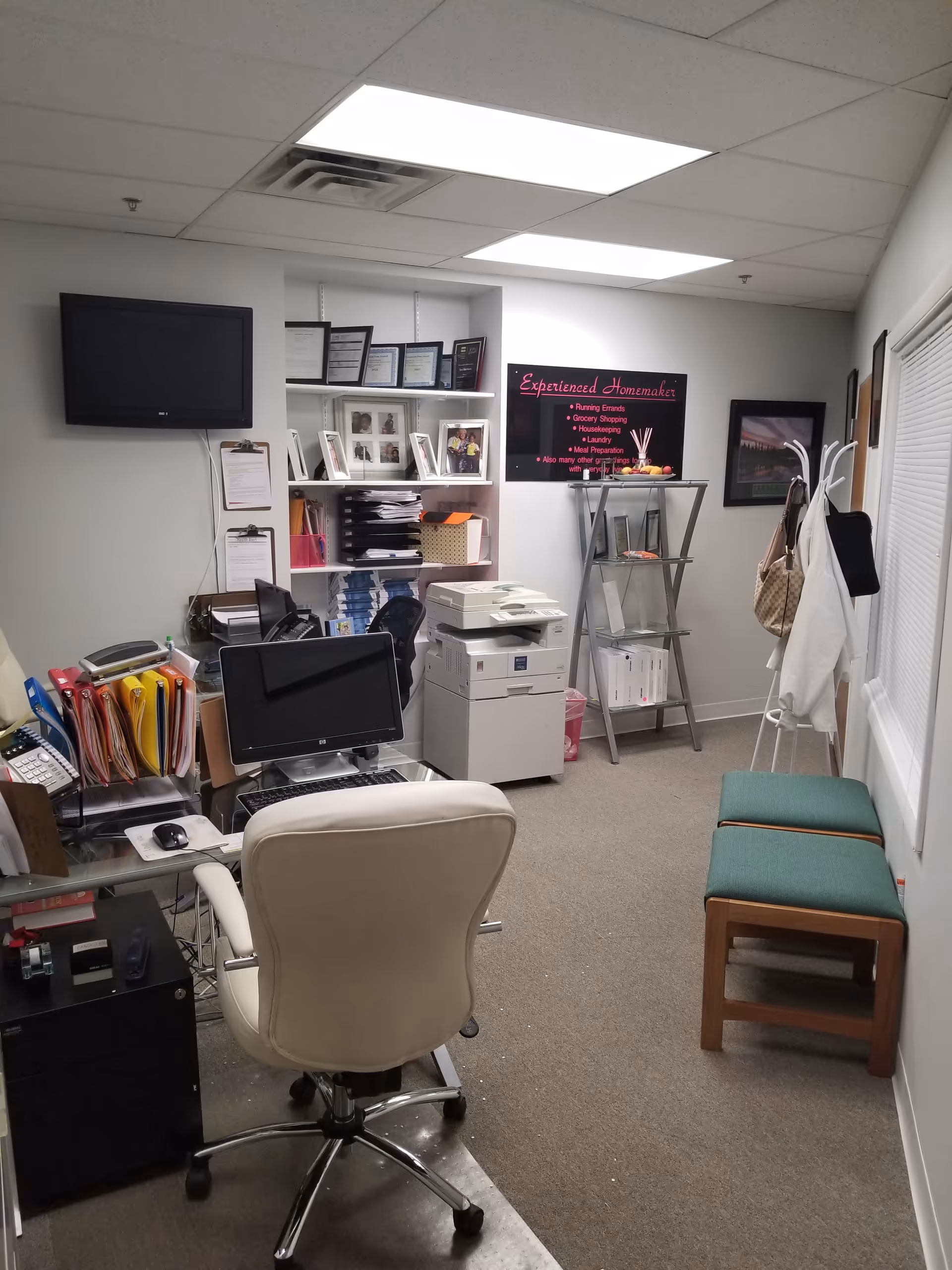 Office workspace with desk, computer, printer, shelves, and coats in a care facility.