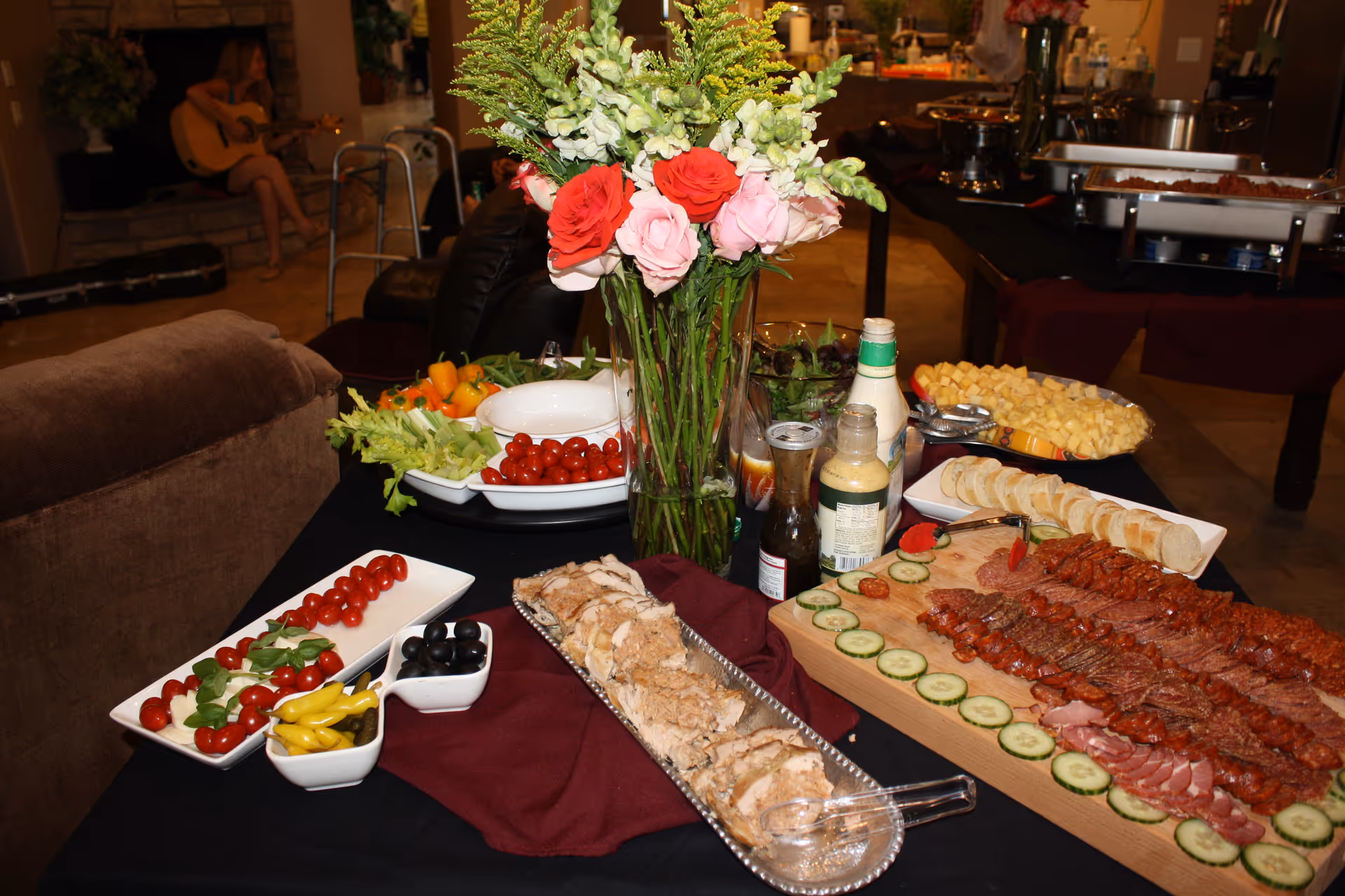 A long buffet table with assorted appetizers, a flower centerpiece, and background residents in dining area.